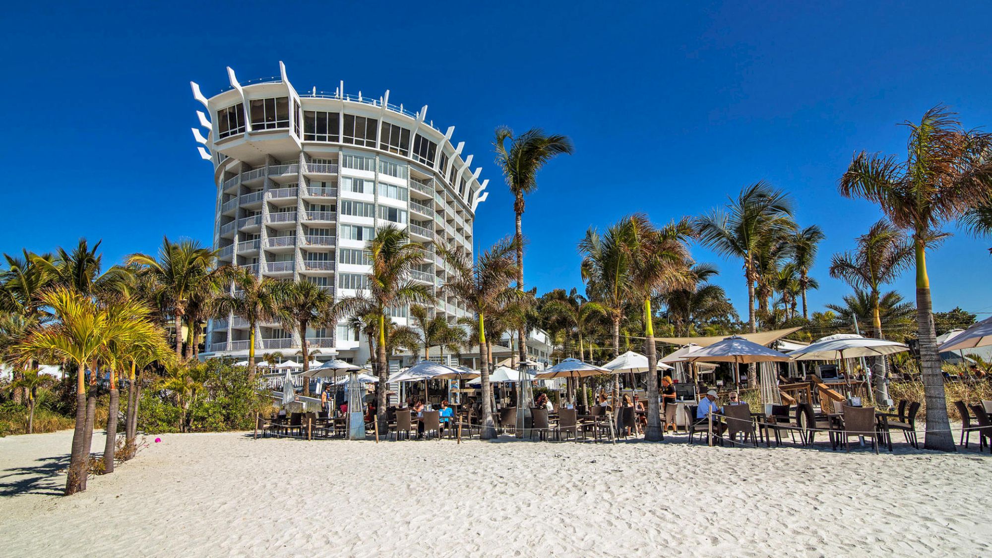 A modern beachfront hotel with white balconies and unique architectural design, surrounded by palm trees and an outdoor seating area with umbrellas on the sand.