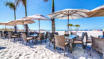 Outdoor beachside dining area with tables and chairs under umbrellas on sandy beach with palm trees and a clear blue sky.