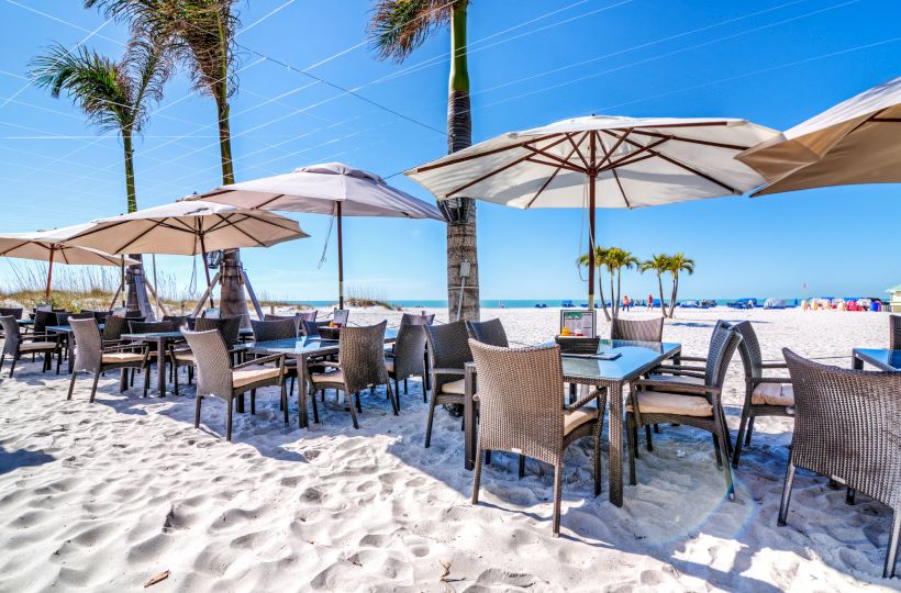 Outdoor beachside dining area with tables and chairs under umbrellas on sandy beach with palm trees and a clear blue sky.