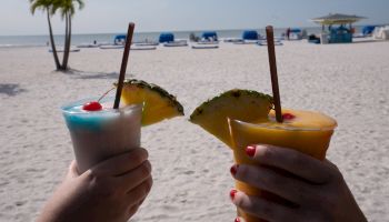 Two hands holding tropical drinks with pineapple slices on a beach with white sand, palm trees, and blue lounge chairs in the background.