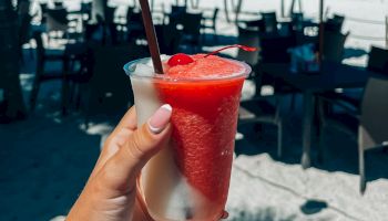 A hand holds a red and white frozen drink with a straw at a beachside venue with blue umbrellas, tables, and chairs, under a clear blue sky.