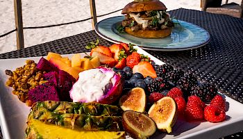 A plate of fresh fruit and a burger on a table by a beach with palm trees and a clear blue sky in the background.