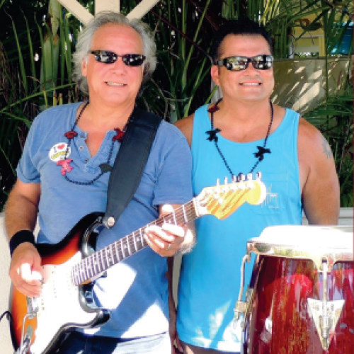 Two individuals wearing sunglasses, one with a guitar and the other next to a drum, are smiling in an outdoor setting.