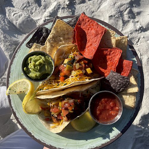 A plate with tacos, red and yellow tortilla chips, guacamole, salsa, and lemon slices is placed on a sandy beach background.