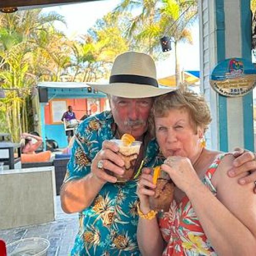 Two people in tropical attire enjoying drinks at an outdoor setting, with palm trees and tropical decor in the background.