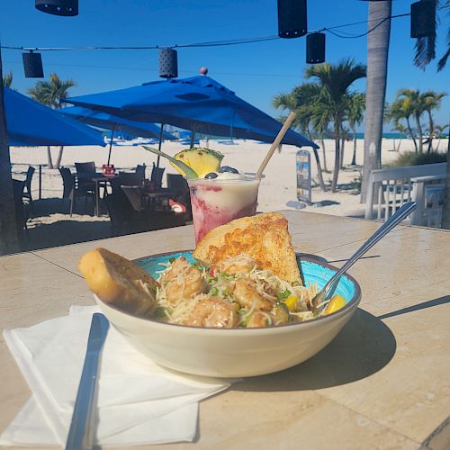 A bowl of food and a tropical drink on a table by the beach with palm trees, umbrellas, and a few tables in the background under a clear blue sky.