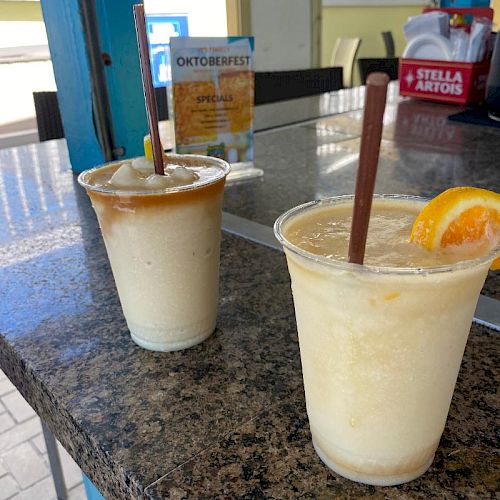 Two frozen drinks with straws on a counter, one garnished with an orange slice. Background shows seating and a sign with an Oktoberfest banner.