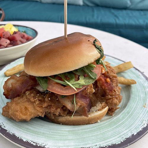 A fried chicken sandwich with lettuce, tomato, and bacon on a plate with fries, alongside a bowl of fruit salad in the background.