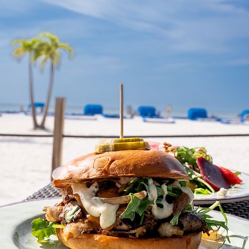 A gourmet burger on a plate with a beach backdrop featuring palm trees, beach chairs, and umbrellas under a clear blue sky.