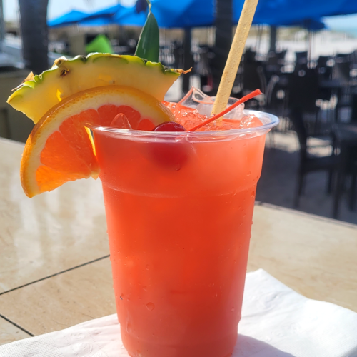 A tropical cocktail garnished with a pineapple slice, orange wedge, and cherry, placed on a napkin at an outdoor seating area by the beach.