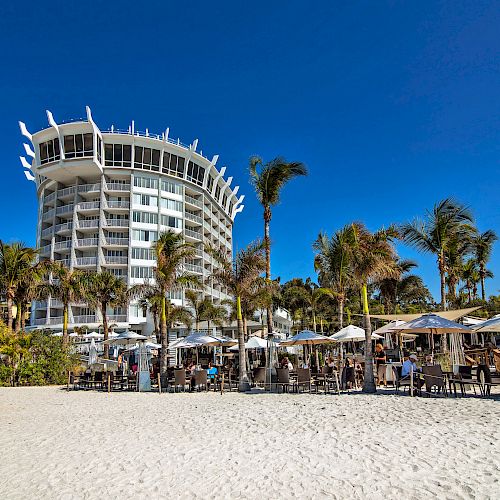 A tall, modern building with unique architecture is surrounded by palm trees, with a sandy beach and outdoor seating in the foreground.