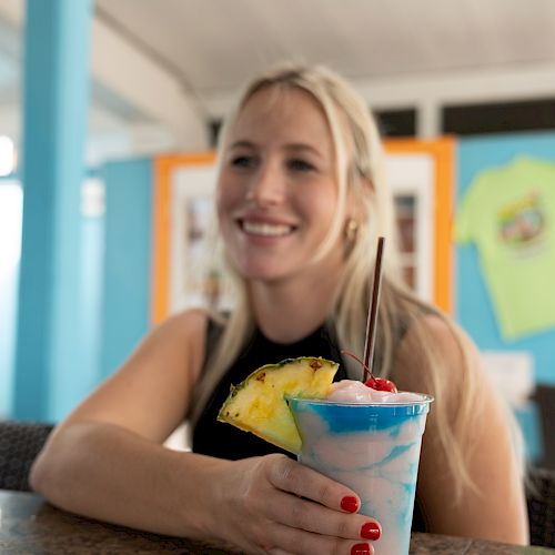 A woman with blonde hair is sitting at a table, holding a colorful drink garnished with a pineapple slice and cherry, in a brightly lit setting.