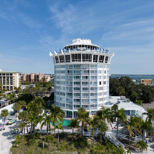 A tall, circular building with multiple floors is surrounded by palm trees near a body of water. It is situated in a scenic, coastal area.