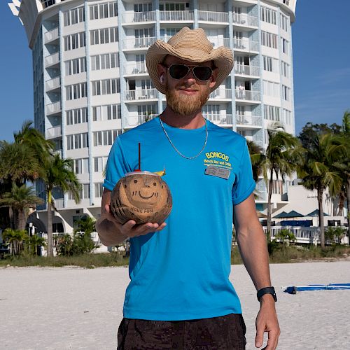 A person in a blue shirt and straw hat stands on a beach, holding a coconut with a face, with a white circular building and palm trees in the background.