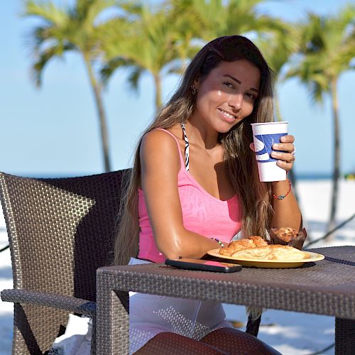 A woman is sitting at an outdoor table on a beach, holding a drink and smiling, with palm trees and clear blue skies in the background.