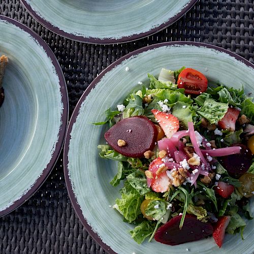 This image shows a plate of fresh salad with various vegetables and a plate with a sandwich cut in half on a woven mat.