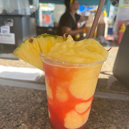 A tropical frozen drink with a swirl of yellow and red, garnished with a pineapple slice and a straw, sits on a counter in an outdoor setting.