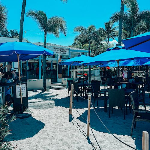 The image shows an outdoor beachside café with tables and chairs under blue umbrellas, surrounded by palm trees, on a bright sunny day.