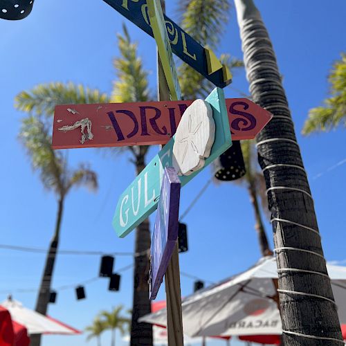 A beach scene with a signpost pointing to various locations like pool, drinks, and gulf, surrounded by palm trees and outdoor seating with umbrellas.