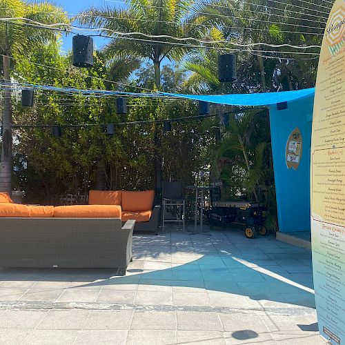 An outdoor seating area with an orange couch, palm trees, blue canopy, and a surfboard menu.