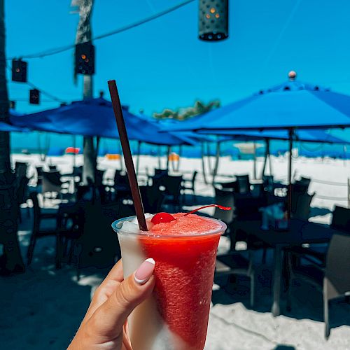 A hand holds a tropical frozen drink with a beach scene in the background; blue umbrellas and tables are visible on a sunny day.