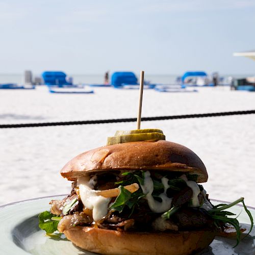 A savory burger topped with greens and cheese, secured by a toothpick, is placed on a plate with a beach in the background.