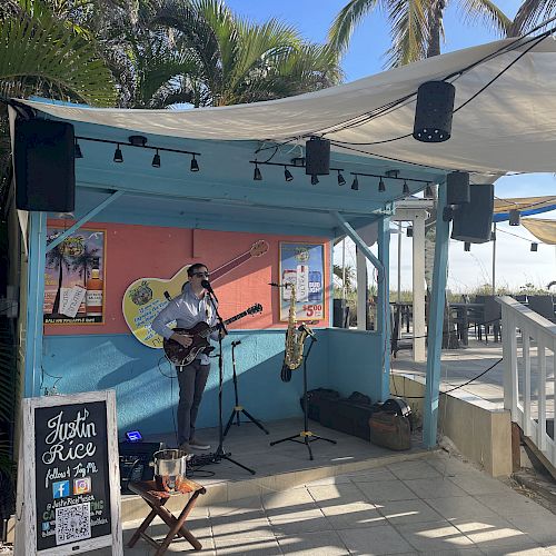 A musician is performing on a small outdoor stage with a guitar. There is a sign in front advertising 