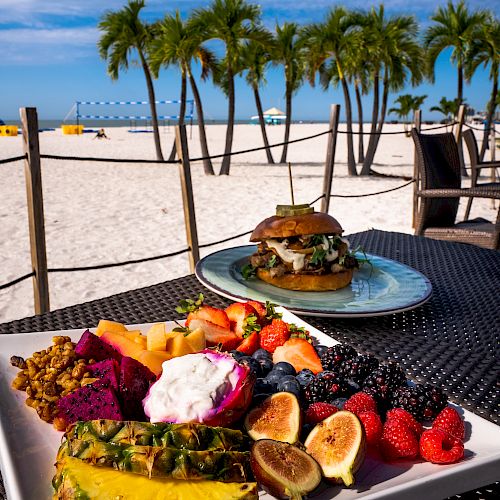 A beachside meal featuring a plate of fresh fruit and a sandwich, with a scenic view of palm trees and a sandy beach in the background.