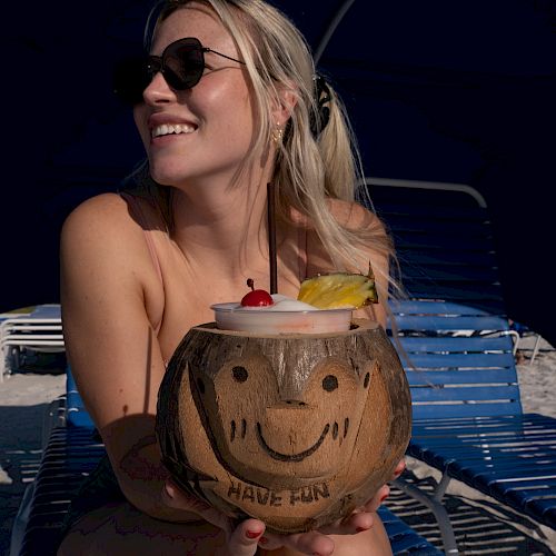 A person is holding a smiling coconut drink with cherries and pineapple on a beach, wearing sunglasses and sitting on a blue lounge chair, ending the sentence.