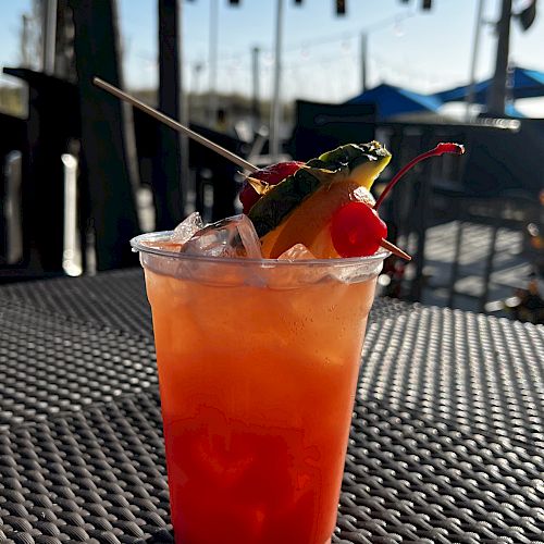 A plastic cup filled with an iced, colorful drink garnished with fruits and a cocktail umbrella, set on a textured outdoor table.