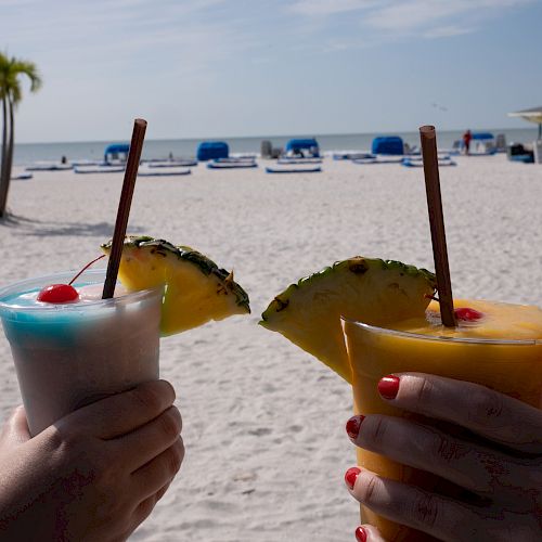 Two hands holding tropical drinks with pineapple garnish and cherries on a beach with white sand, palm trees, and blue beach chairs.