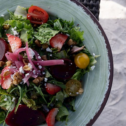 A salad on a green plate with beets, strawberries, tomatoes, mixed greens, nuts, and cheese, placed next to a sandy surface.