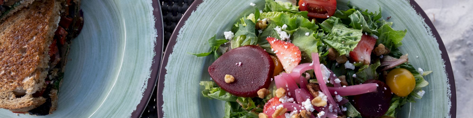 A plate with a fresh salad featuring beets, strawberries, cherry tomatoes, and greens, and another plate with a sandwich split in half.