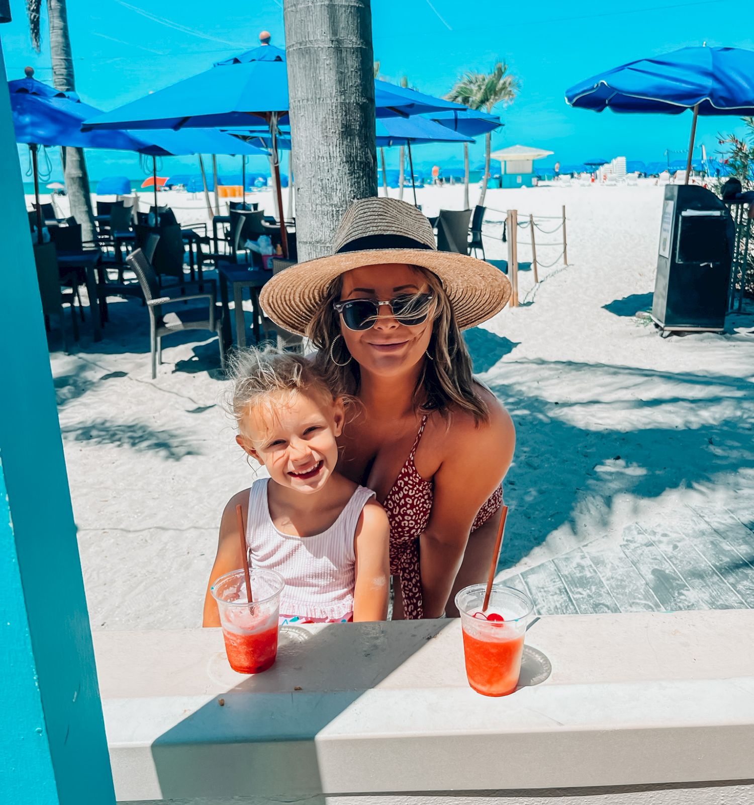 A woman and a child are at a beachside café with drinks, under blue umbrellas, with palm trees and the ocean in the background.