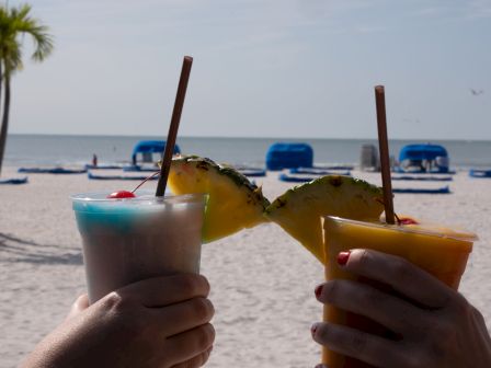 Two people holding tropical drinks with fruit garnishes on a beach with blue lounge chairs and a palm tree in the background, sky clear.