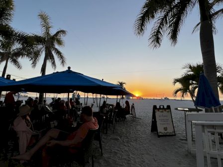 People are dining under blue umbrellas on a beach at sunset, with palm trees and a signboard in the background.