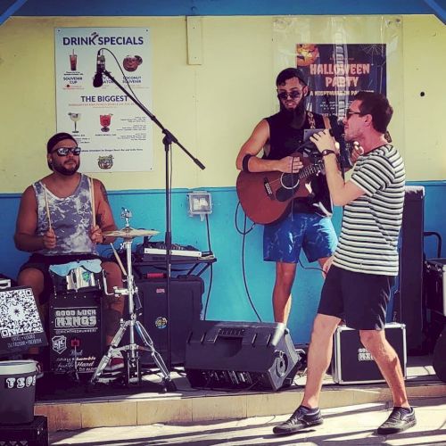 Three musicians are performing on a small stage; one on drums, one with a guitar, and one singing to a microphone while holding it.