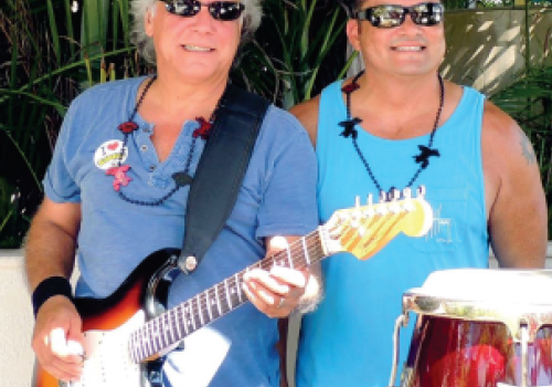 Two people wearing sunglasses, standing outdoors; one plays an electric guitar, the other stands beside conga drums, both smiling.