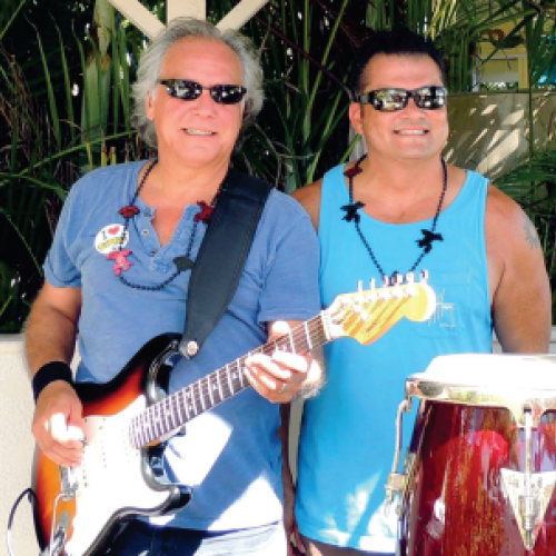 Two people wearing sunglasses, standing outdoors; one plays an electric guitar, the other stands beside conga drums, both smiling.