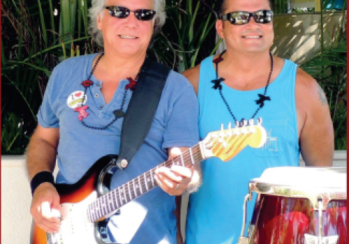 Two men are smiling and standing together outdoors; one is holding an electric guitar, and the other is standing next to congas.