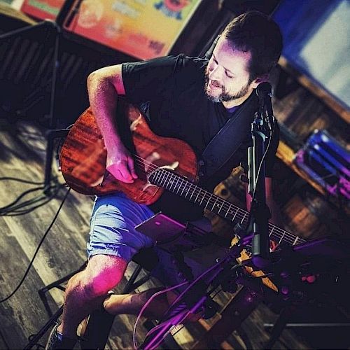 A person is sitting and playing an acoustic guitar in a dimly lit room, with music equipment and a microphone nearby.