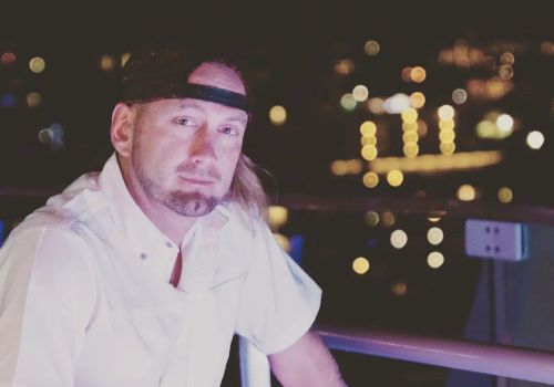 A person wearing a white shirt and a cap is sitting outdoors at night with city lights blurred in the background.