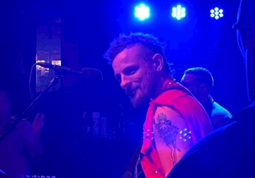 A musician with a red sleeveless jacket and tattoos plays guitar on stage under blue lights, with other band members in the background.