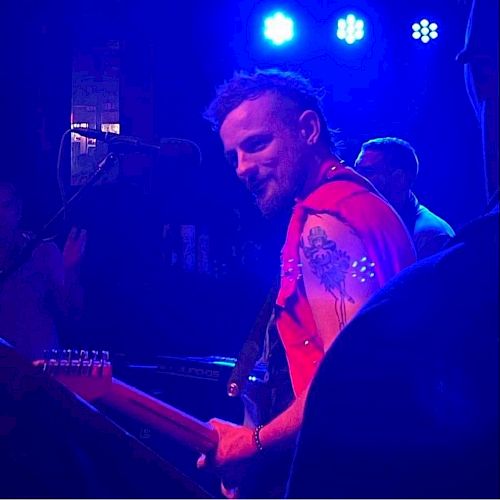 A musician with a red sleeveless jacket and tattoos plays guitar on stage under blue lights, with other band members in the background.