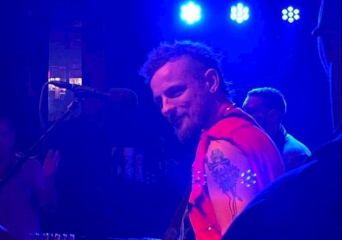 A musician with a beard and a red vest plays the guitar on stage under blue lights.