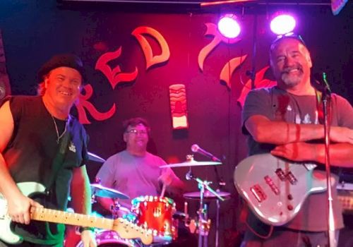 Three musicians with guitars and drums perform on a dimly lit stage, smiling, under neon 