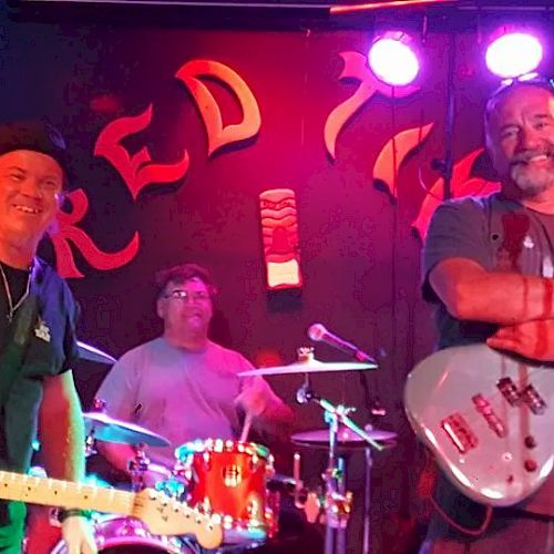 Three musicians with guitars and drums perform on a dimly lit stage, smiling, under neon 