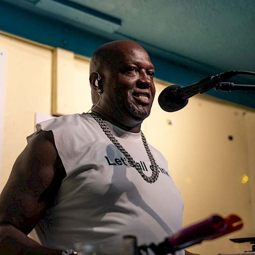 A man stands at a microphone in a room, possibly a bar or restaurant, with a menu on the wall in the background.