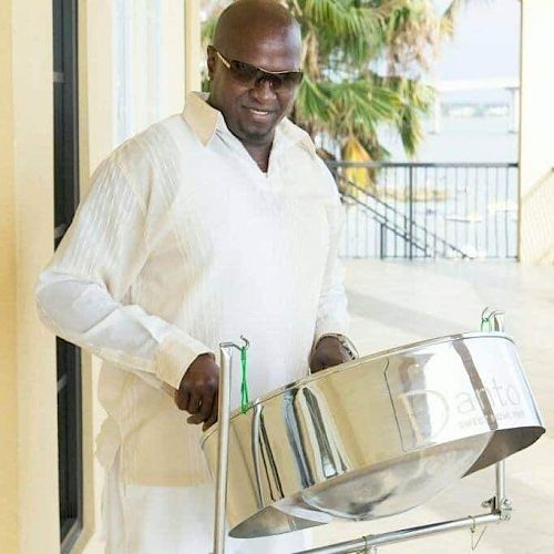 A person wearing sunglasses and a white outfit is playing a steel drum outdoors, with palm trees and railings in the background.