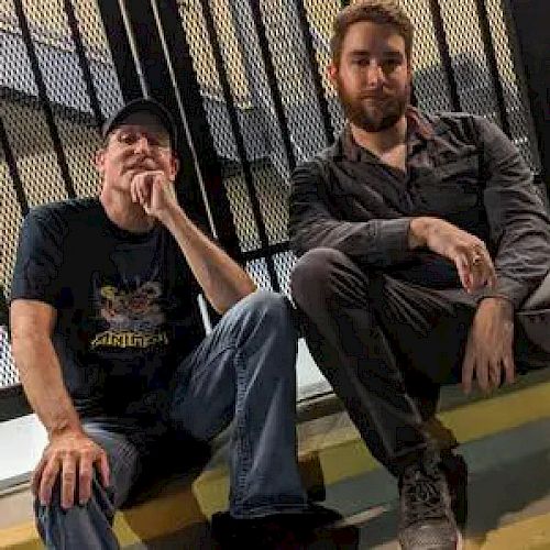 Two men are seated on steps in front of a metal fence, one wearing a black shirt and cap, the other in a gray shirt, with relaxed expressions.